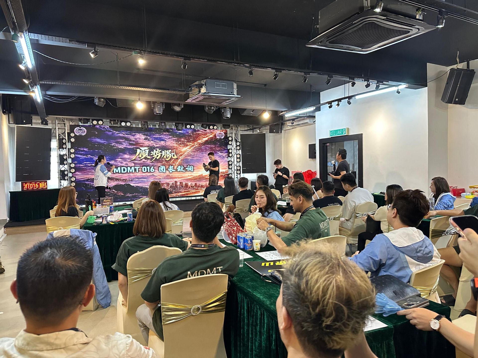 Corporate team gathering with members in team shirts at banquet tables and a leader speaking on stage at Dreamlive Event Space Puchong